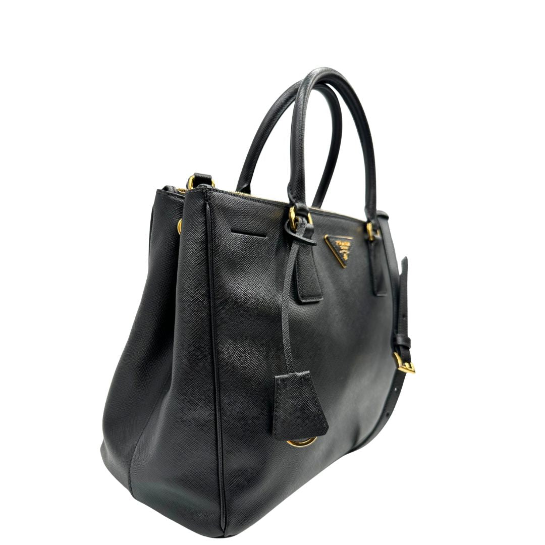 Laterale sinistro di una borsa Prada in pelle saffiano nera con parti metalliche dorate, munita di doppi manici stondati, tracolla amovibile e regolabile ed una clochette portachiavi. Completa di dustbag