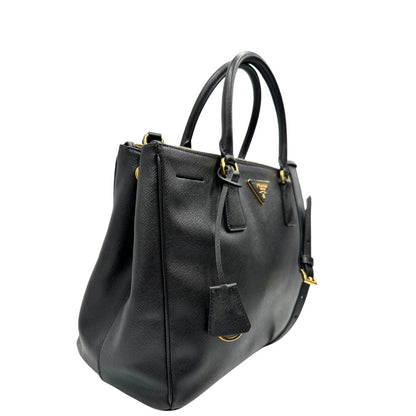 Laterale sinistro di una borsa Prada in pelle saffiano nera con parti metalliche dorate, munita di doppi manici stondati, tracolla amovibile e regolabile ed una clochette portachiavi. Completa di dustbag