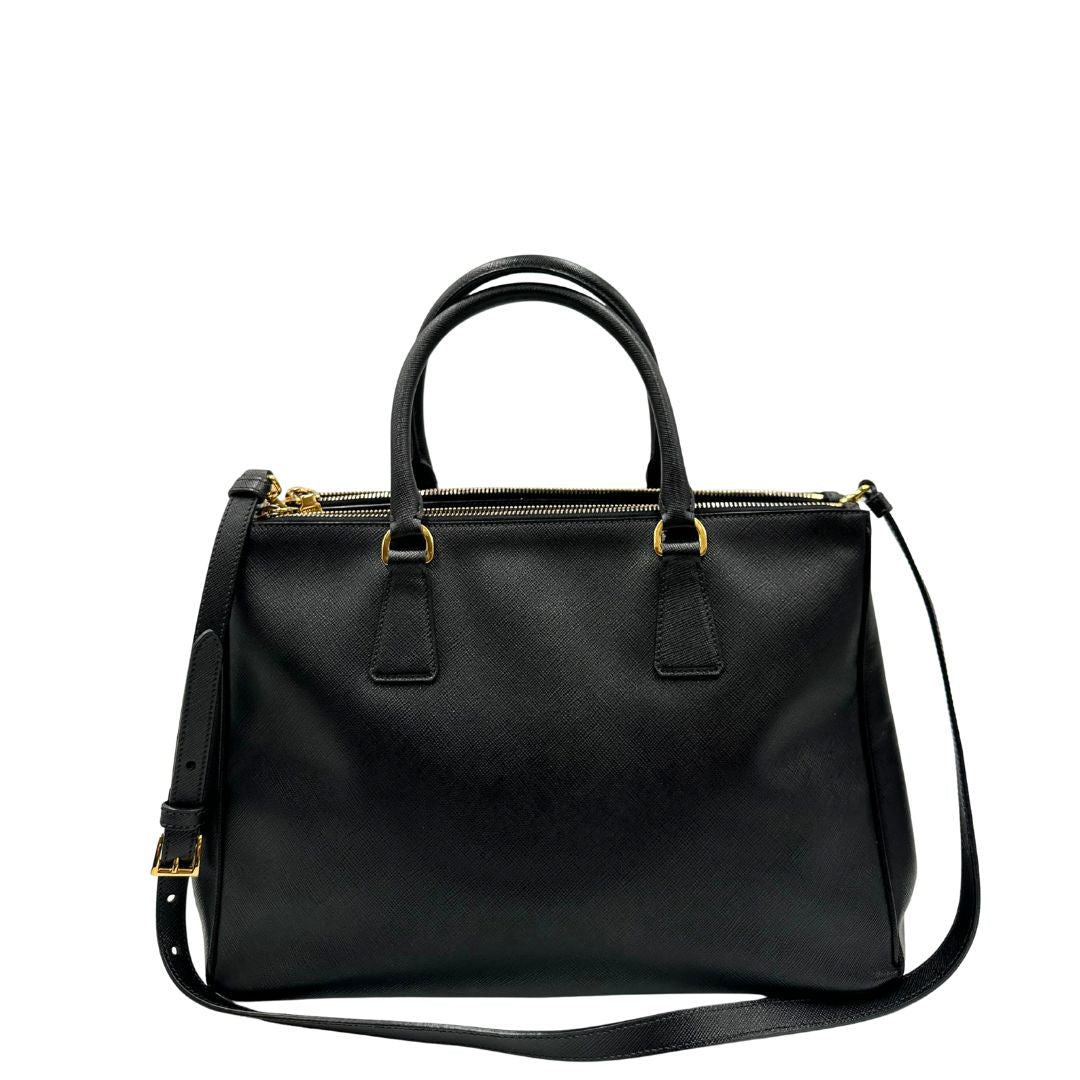 Retro di una borsa Prada in pelle saffiano nera con parti metalliche dorate, munita di doppi manici stondati, tracolla amovibile e regolabile ed una clochette portachiavi. Completa di dustbag