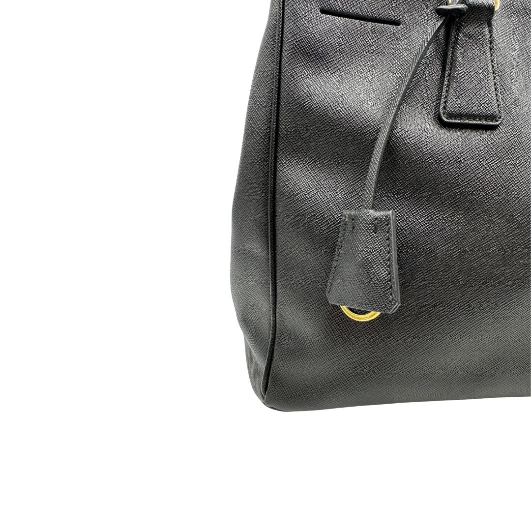 Clochette portachiavi di una borsa Prada in pelle saffiano nera con parti metalliche dorate, munita di doppi manici stondati, tracolla amovibile e regolabile. Completa di dustbag.