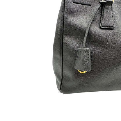 Clochette portachiavi di una borsa Prada in pelle saffiano nera con parti metalliche dorate, munita di doppi manici stondati, tracolla amovibile e regolabile. Completa di dustbag.