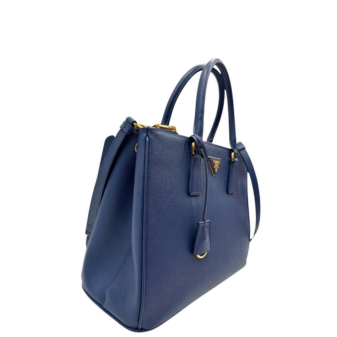 lato Borsa Prada in pelle saffiano blu con parti metalliche dorate; munita di doppi manici stondati, una tracolla amovibile e regolabile e impreziosita da un portachiavi, usata, originale, ottime condizioni. 