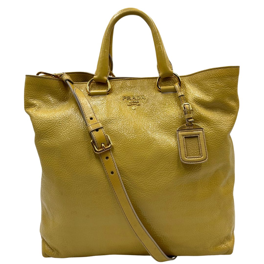 Front di una Borsa Prada in pelle lucida color mustard con parti metalliche dorate; munita di un accessorio porta address e doppi manici stondati. Indossabile a mano oppure a tracolla. Originale, usata, di lusso, in ottime condizioni