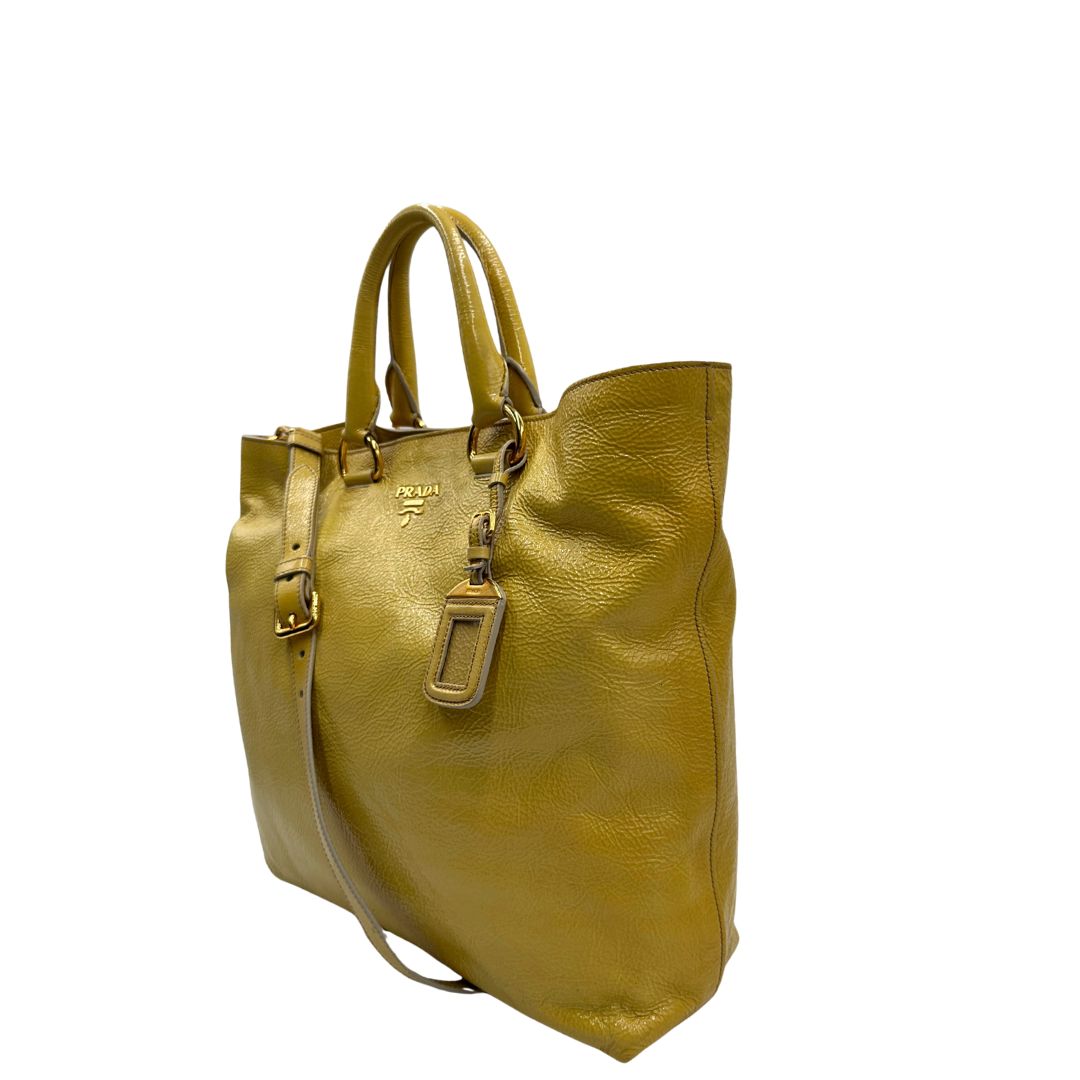 Lato sinistro di una Borsa Prada in pelle lucida color mustard con parti metalliche dorate; munita di un accessorio porta address e doppi manici stondati. Indossabile a mano oppure a tracolla. Originale, usata, di lusso, in ottime condizioni