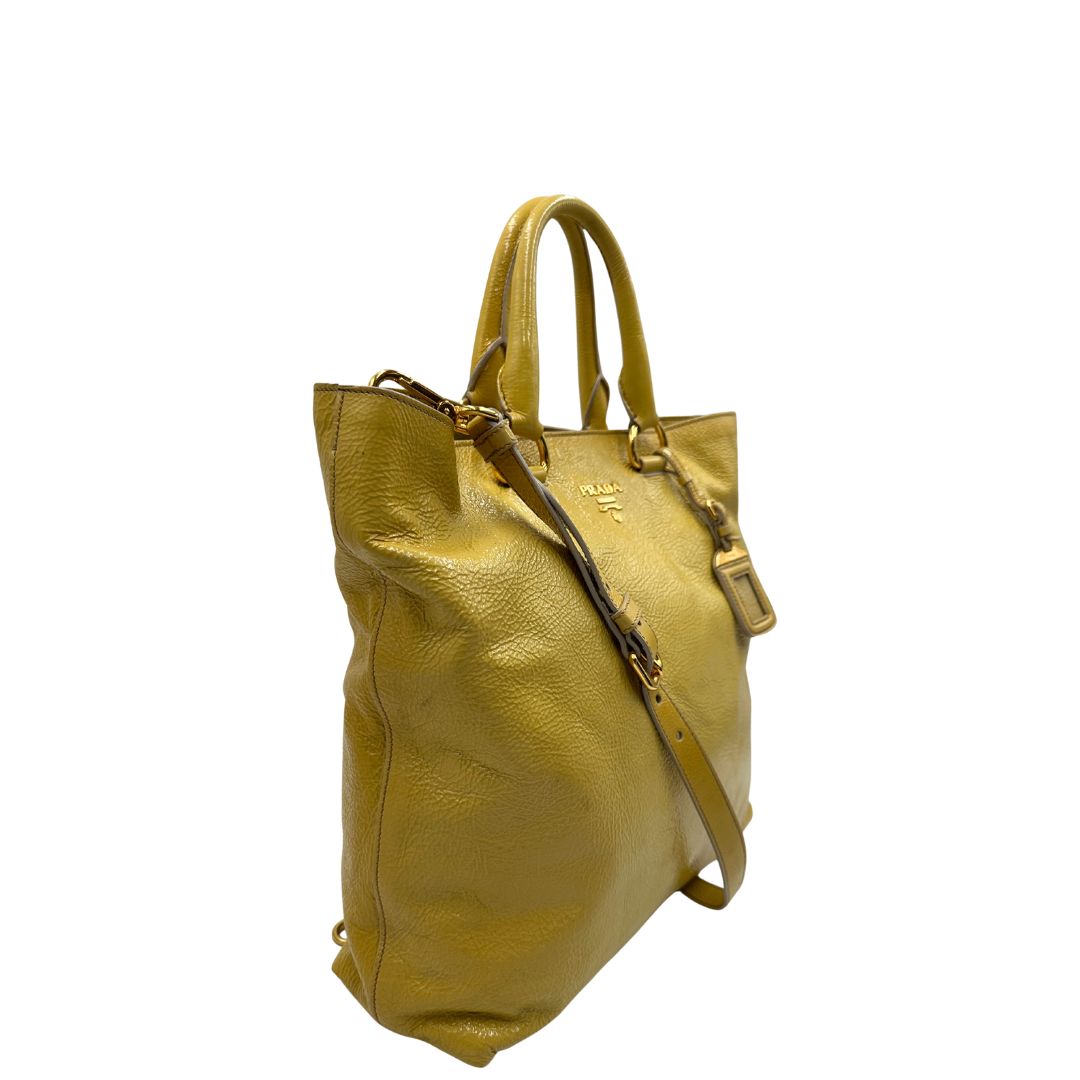 Lato destro di una Borsa Prada in pelle lucida color mustard con parti metalliche dorate; munita di un accessorio porta address e doppi manici stondati. Indossabile a mano oppure a tracolla. Originale, usata, di lusso, in ottime condizioni