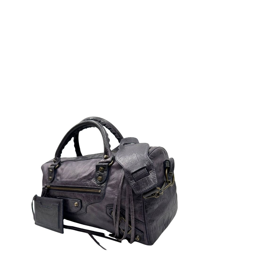 Lato sinistro di una Borsa Balenciaga in pelle viola scuro effetto invecchiato con parti metalliche ottone anticato, munita di doppi manici con dettaglio intreccio, e una tracolla amovibile e regolabile con spallaccio. Completa di dustbag. Originale, usata, di lusso, in ottime condizioni.