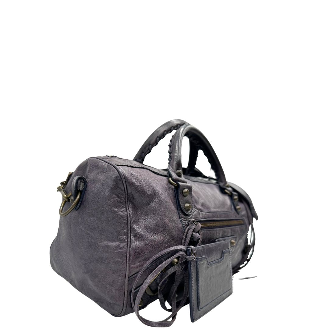 Lato destro di una Borsa Balenciaga in pelle viola scuro effetto invecchiato con parti metalliche ottone anticato, munita di doppi manici con dettaglio intreccio, e una tracolla amovibile e regolabile con spallaccio. Completa di dustbag. Originale, usata, di lusso, in ottime condizioni.