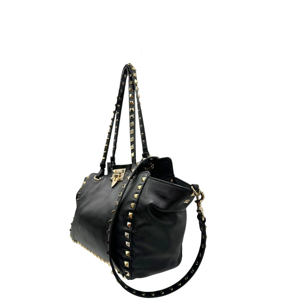 Lato sinistro di una Borsa Valentino Garavani in pelle nera con parti metalliche dorate; impreziosita da borchie piramidali e munita di doppi manici e una tracolla amovibile. Completa di dustbag e cartellino. Originale, usata, di lusso, in ottime condizioni.