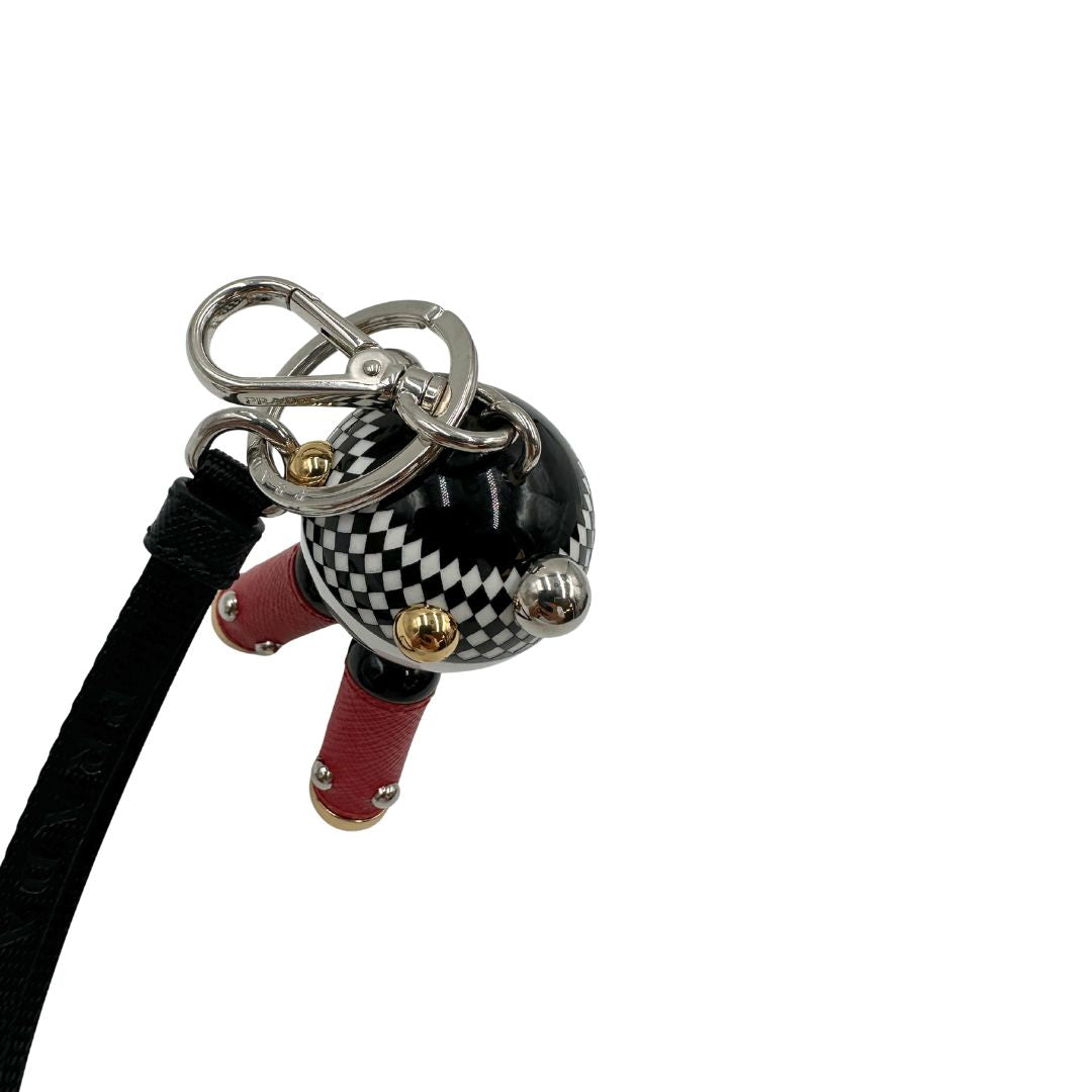 Charm Prada presenta una testa a sfera con un motivo a scacchi bianchi e neri e dettagli metallici color oro, le gambe sono realizzate in pelle saffiano rossa. Originale, usato, di lusso, in condizioni eccellenti.