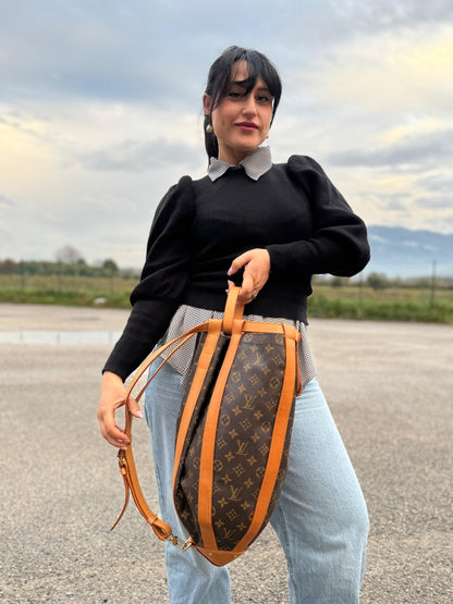 Ragazza che mostra una borsa Louis Vuitton limited edition Romeo Gigli, in canvas Monogram e pelle naturale. Struttura cilindrica con rinforzi in pelle e finiture metalliche dorate. Chiusura superiore con coulisse. Spallaccio in pelle regolabile. Dotata di dustbag e manico amovibile. Indossabile a spalla.
