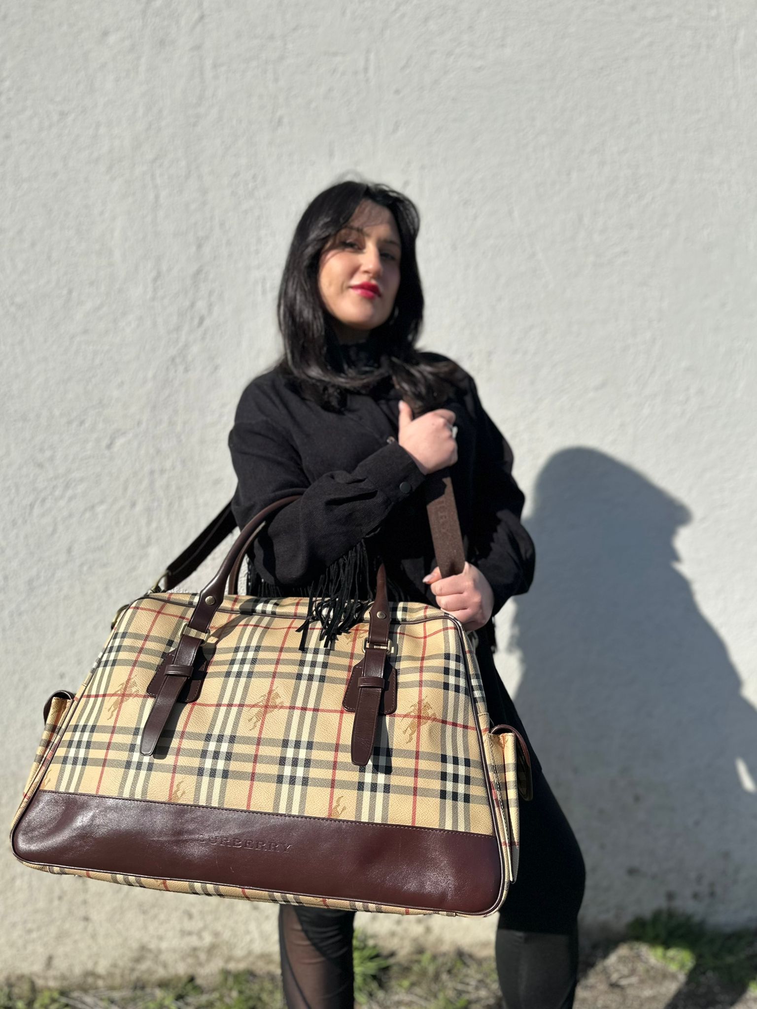 Ragazza che indossa un borsone Burberry in canvas trama tartan con finiture in pelle marrone testa di moro e parti metalliche dorate; munito di doppi manici stondati e una tracolla amovibile e regolabile.