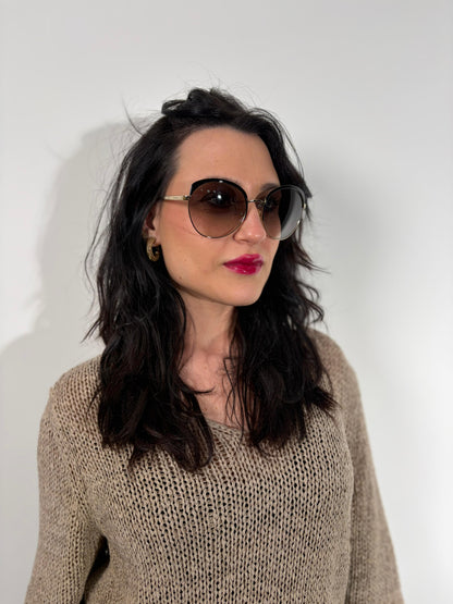 Ragazza che indossa un paio di occhiali da sole Prada caratterizzati da una montatura nera lucida con lenti sfumate grigie, asticelle maculate e hardware dorato, usati, originali, second hand, in ottime condizioni.
